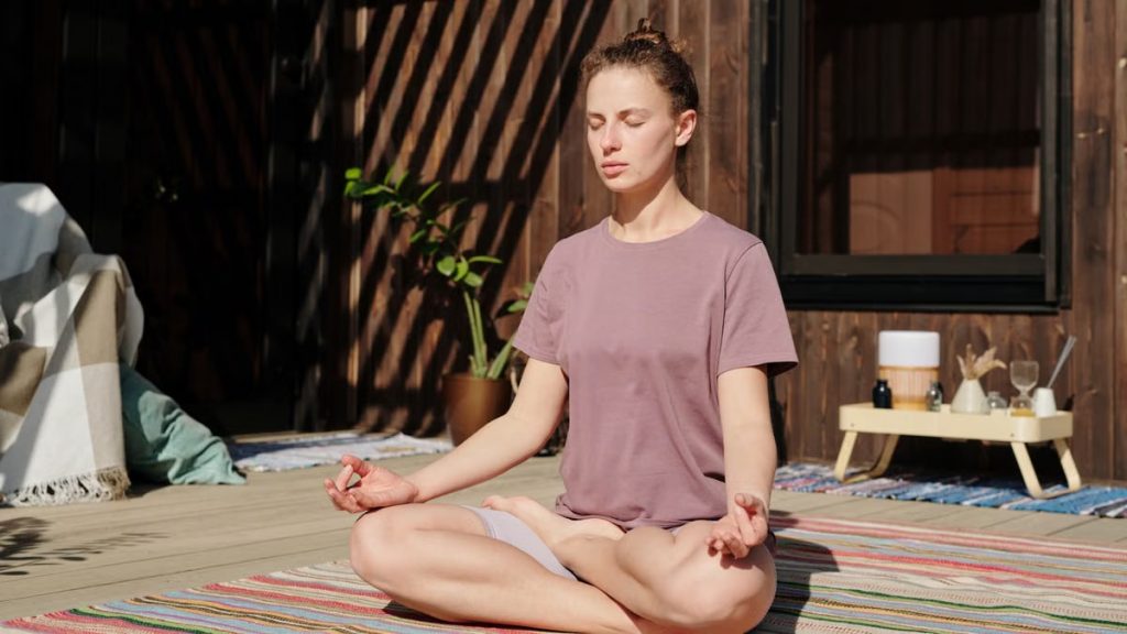 Woman practicing meditation in lotus pose on a sunny outdoor deck.